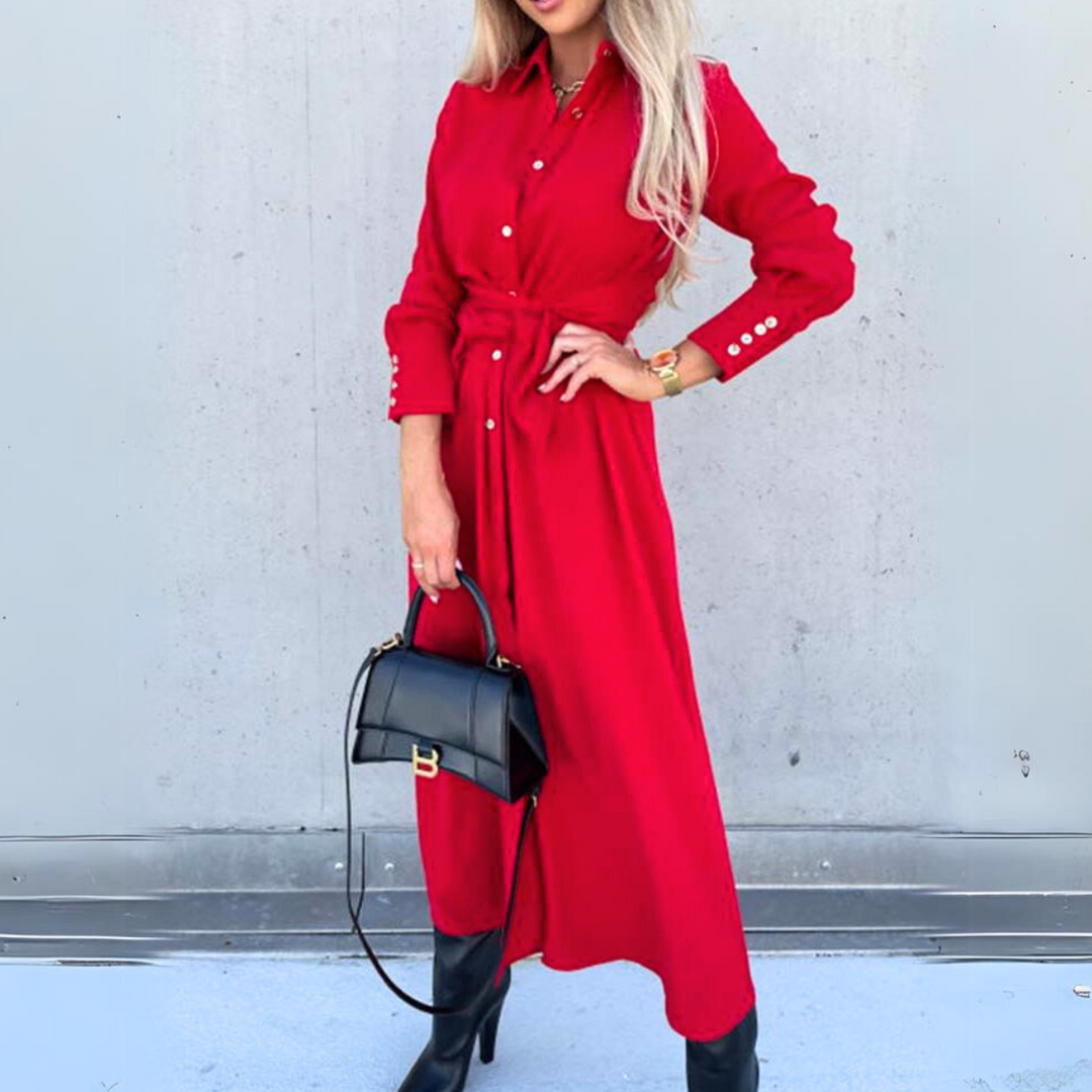 A woman in a long-sleeve, button-down red wrap dress with a waist tie stands against a gray wall, holding a black handbag and wearing black knee-high boots. Her face is not visible, giving the look an effortless high fashion vibe.
