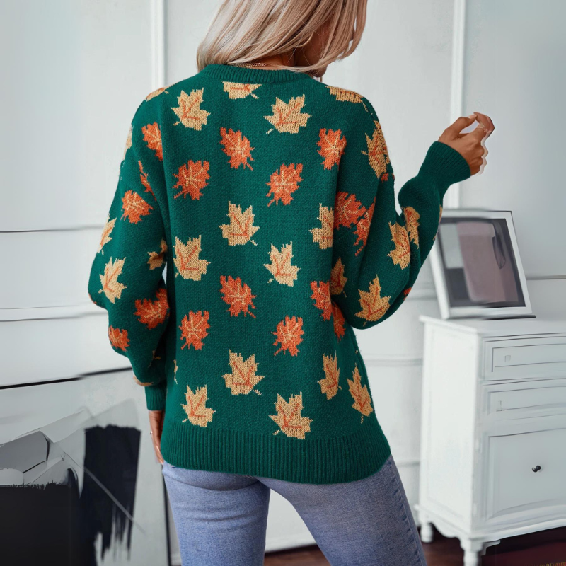 A person with long blonde hair wears a comfortable green sweater featuring a maple leaf pattern in orange and yellow. They are standing indoors, facing away, in a room with white walls and furniture.