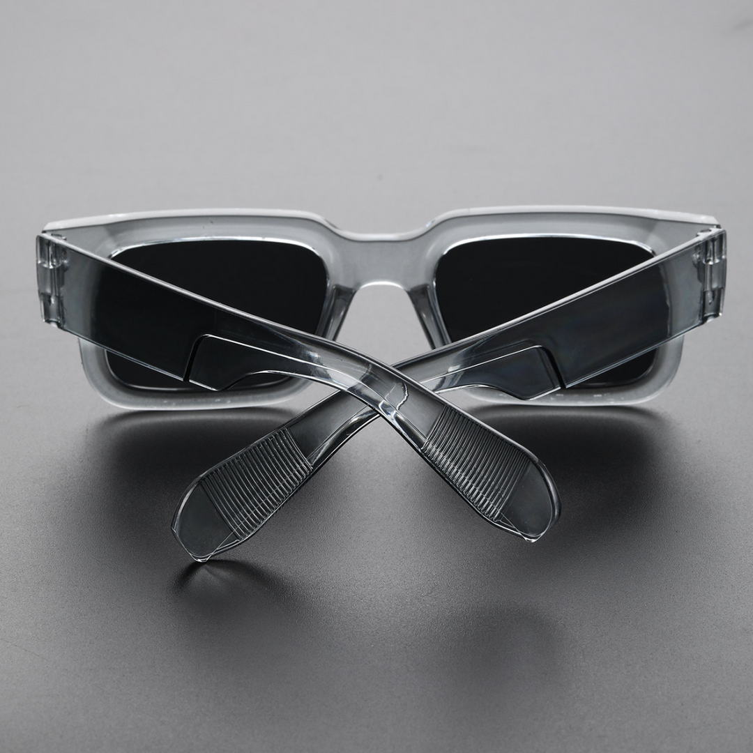 A pair of lightweight sunglasses with bold angular frames and dark lenses is placed upside down on a gray surface, with the temples crossed in front of the lenses.