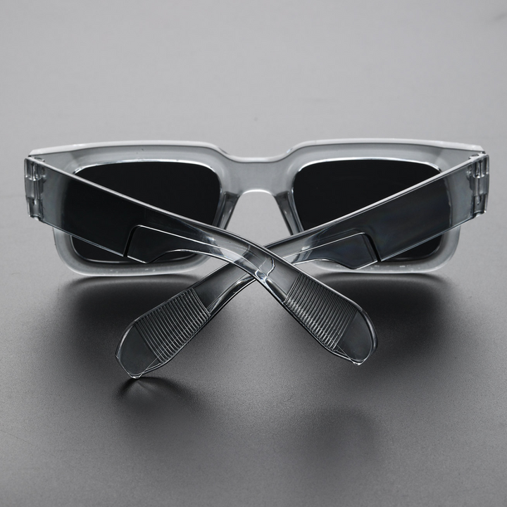 A pair of lightweight sunglasses with bold angular frames and dark lenses is placed upside down on a gray surface, with the temples crossed in front of the lenses.