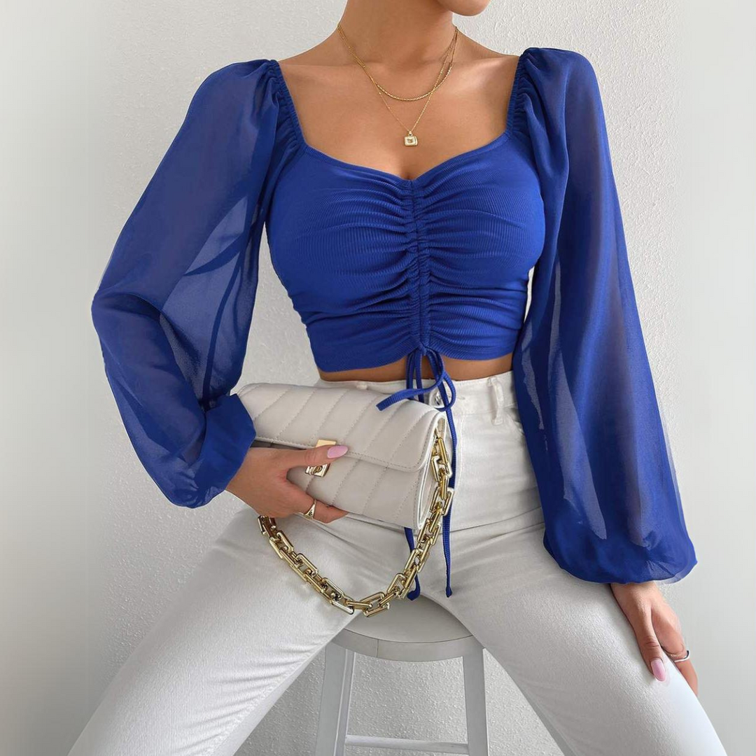 Woman wearing a royal blue ruched crop top with a flirty neckline and sheer long sleeves, white high-waisted pants, gold necklaces, and holding a white quilted purse with a gold chain while sitting on a white stool.