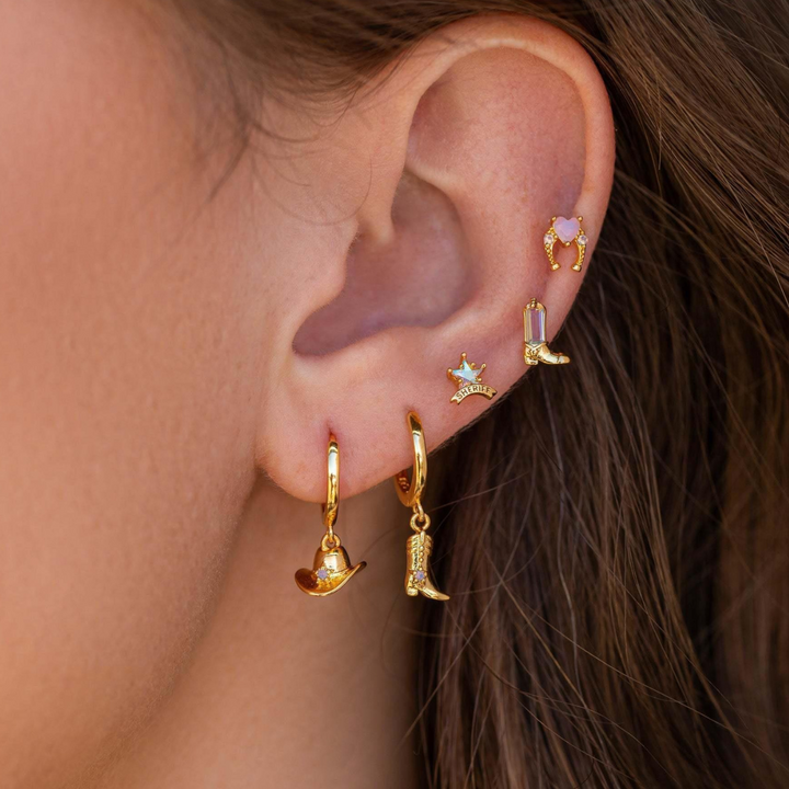A close-up of a persons ear with multiple gold statement earrings, including a cowboy hat, cowboy boot, crown, gem, and shield shapes, set against brown hair and light skin.