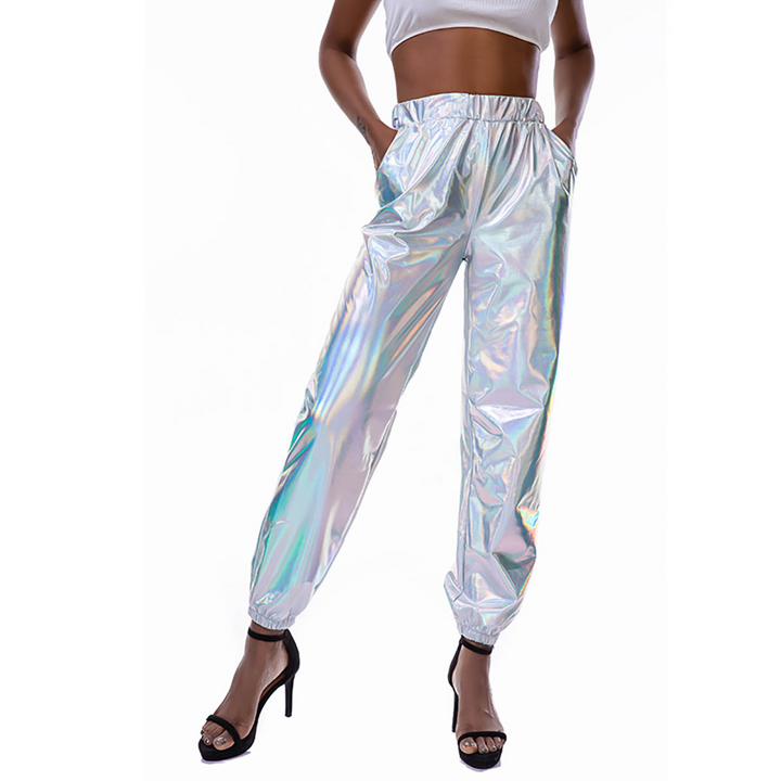 A woman wearing shiny silver jogger pants with a holographic finish and an elastic high waist, a white crop top, and black high-heeled sandals, poses with her hands in her pockets against a white background.