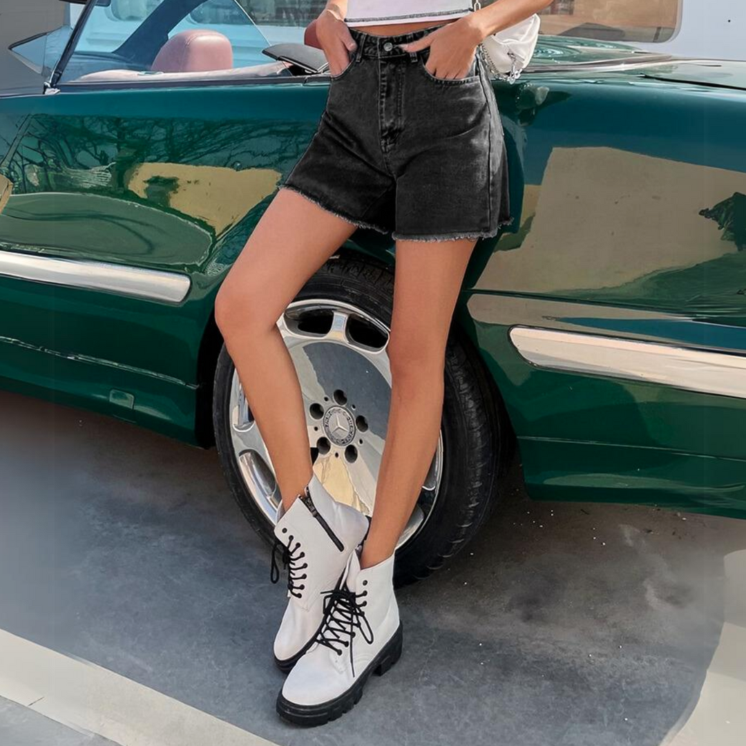 A person wearing high-waist black wash denim streetwear shorts, a light top, and white lace-up boots stands next to a shiny green car with chrome wheels on a sunny day.