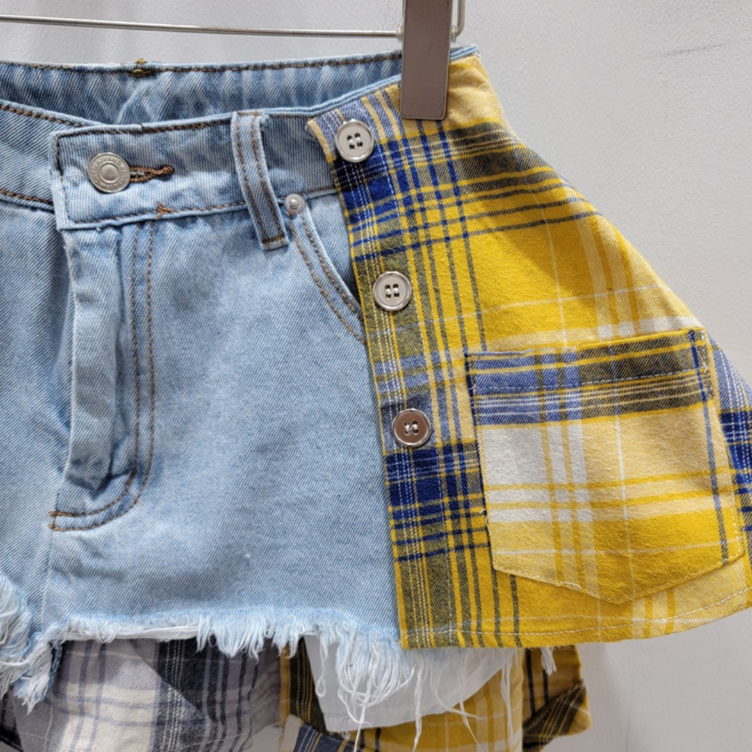 A close-up of a patchwork skirt blending light blue denim with frayed edges and yellow plaid fabric with buttons and a pocket, hanging on a hanger against a white background—a statement piece for eclectic fashion lovers.