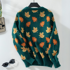 A comfortable sweater in green, featuring an orange and yellow maple leaf pattern, hangs on a black hanger against a white wall. Sunglasses and a small decorative item are nearby.