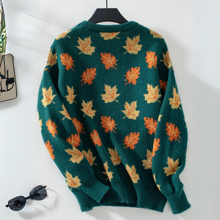 A comfortable sweater in green, featuring an orange and yellow maple leaf pattern, hangs on a black hanger against a white wall. Sunglasses and a small decorative item are nearby.