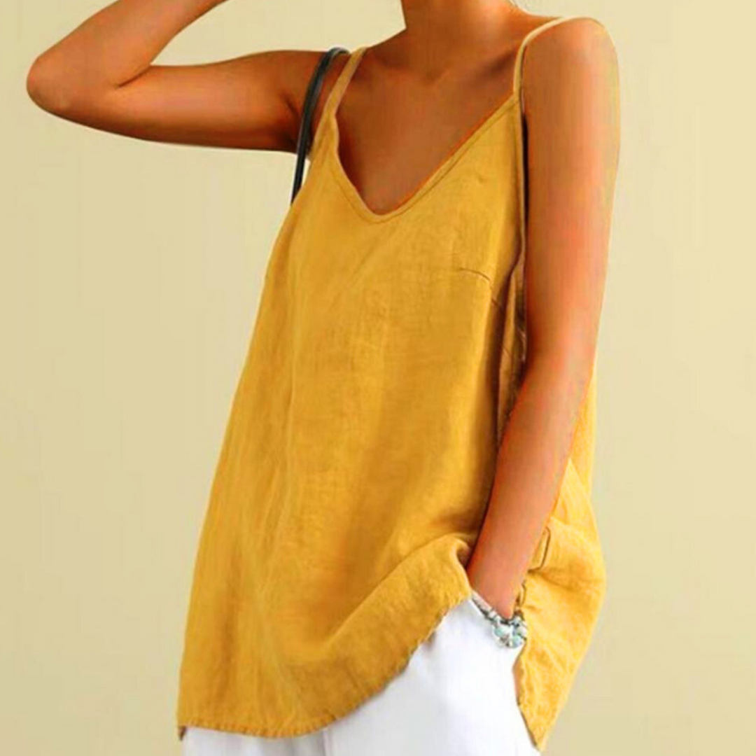 A person wearing a loose, sleeveless mustard yellow urban-chic camisole with spaghetti straps and white pants, standing against a plain light background. One hand is on their hip and the other is resting on their head.