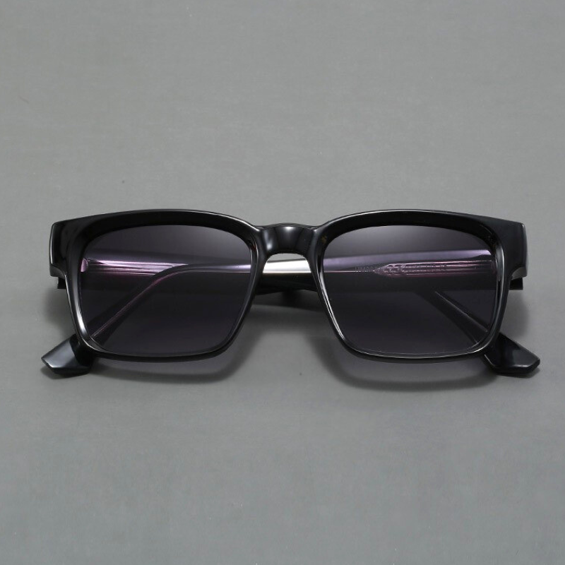 A pair of black rectangular sunglasses with modern design and dark lenses is displayed on a plain gray surface, viewed from the front.