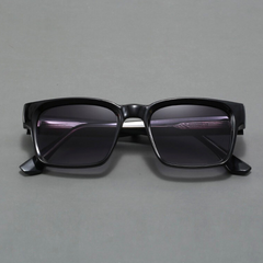 A pair of black rectangular sunglasses with modern design and dark lenses is displayed on a plain gray surface, viewed from the front.