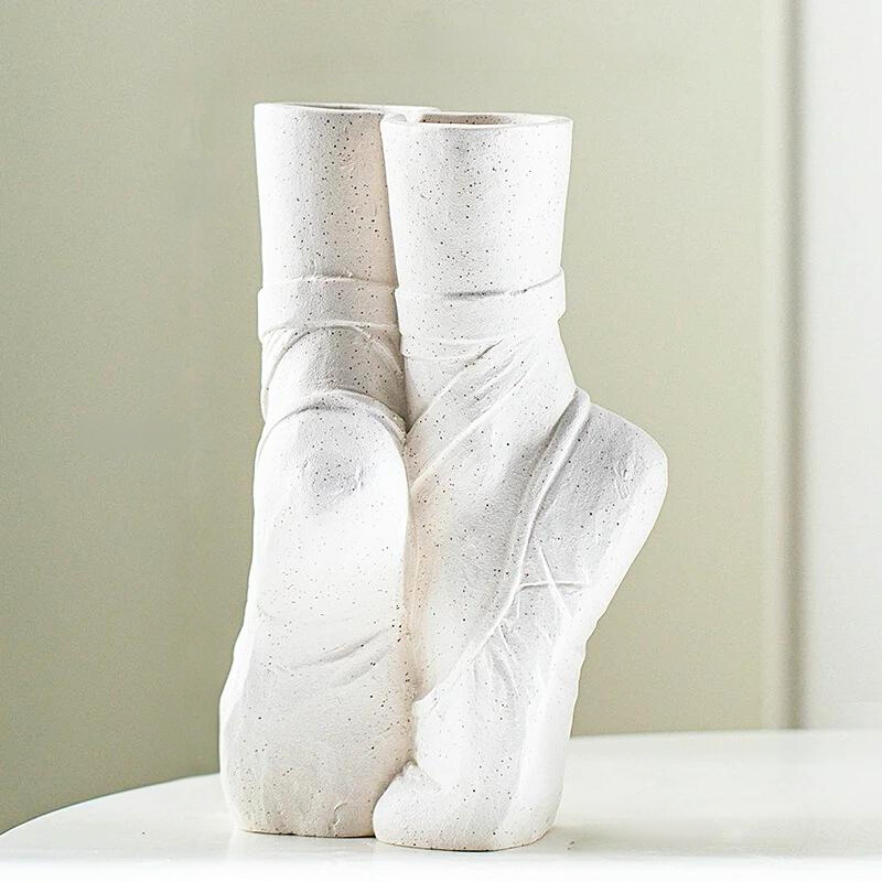 A white ceramic vase resembling two ballet slippers, posed en pointe, sits on a smooth white surface against a light, blurred background—perfect for contemporary chic décor.