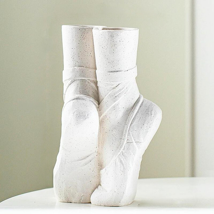 A white ceramic vase resembling two ballet slippers, posed en pointe, sits on a smooth white surface against a light, blurred background—perfect for contemporary chic décor.