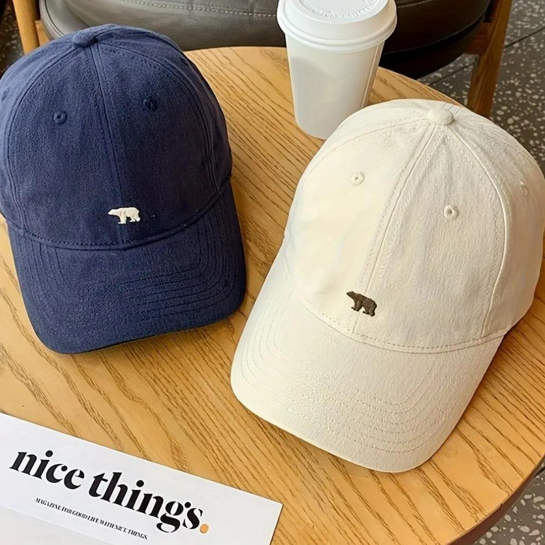 Two premium cotton baseball caps, one navy blue and one cream, each with a small bear logo on the front, rest on a wooden table beside a white coffee cup and a magazine titled nice things—perfect for stylish sun protection.