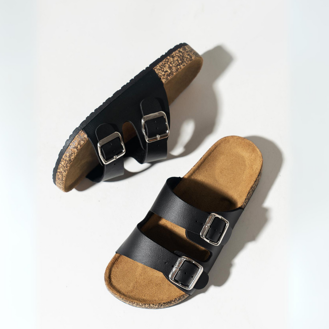 A pair of black double-buckle sandals with premium leather straps and ergonomic cork footbed, positioned on a white background—one sandal lying flat, the other turned on its side.