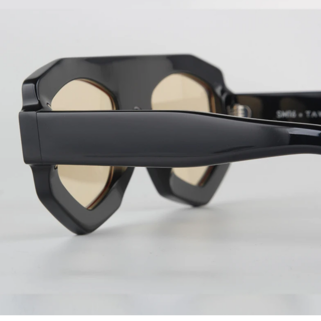 Close-up of black sunglasses with thick, geometric acetate frames and light brown tinted lenses, offering UV400 protection, shown from the side resting on a white surface.