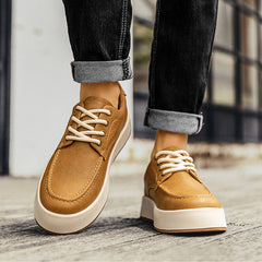 A person wearing rolled-up black jeans and tan men's leather casual shoes with white soles, walking on a wooden surface near a building with large windows.