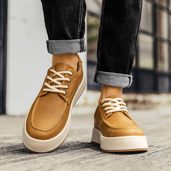 A person wearing rolled-up black jeans and tan men's leather casual shoes with white soles, walking on a wooden surface near a building with large windows.