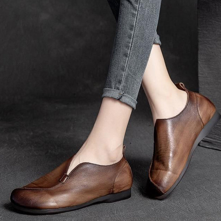 A person wearing brown ankle-high shoes made of supple leather with black soles and rolled-up gray jeans sits against a dark background, with one foot resting on its side.