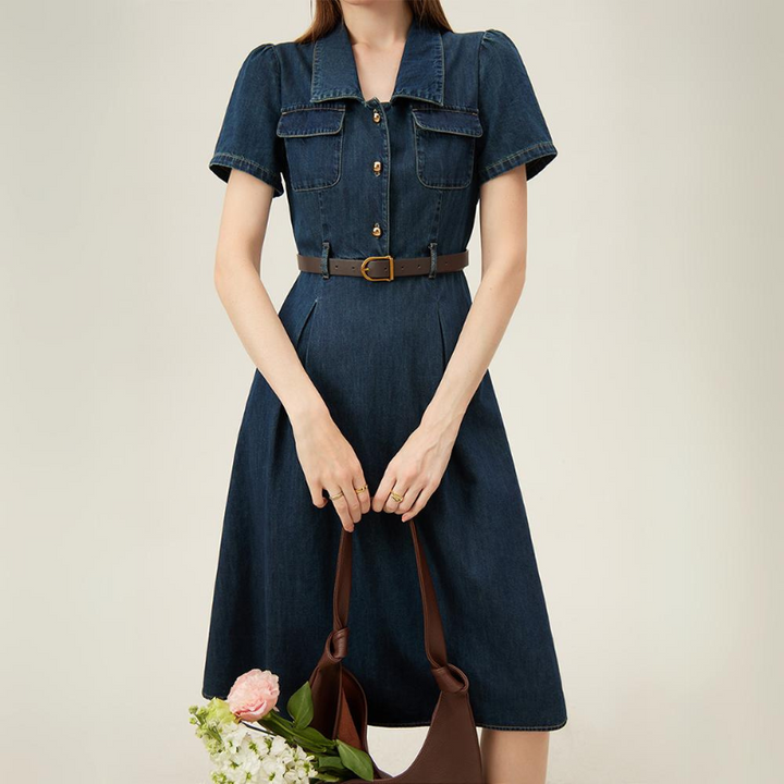 A person in a short-sleeved, belted denim dress stands against a plain background, holding a brown bag with pink and white flowers—an effortless choice for chic summer wear. Their head is not visible in the image.