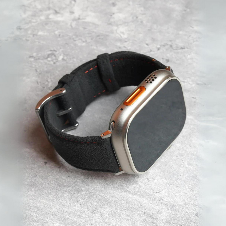 A modern smartwatch with a square face, silver casing, orange button, and a black fabric strap with a stainless steel buckle rests on a light gray, textured surface.
