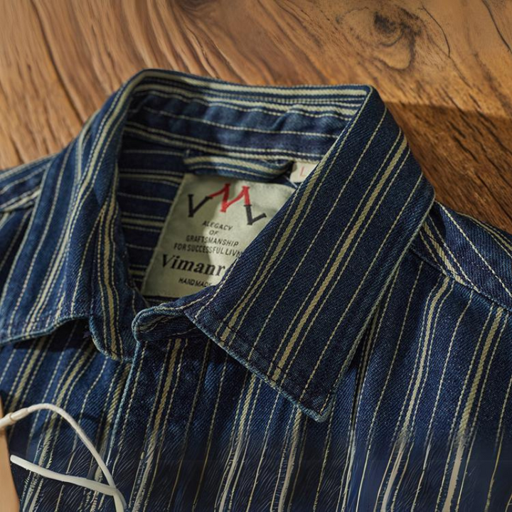 A close-up of a blue vertical striped collared shirt with white and yellow lines, neatly folded on a wooden surface. The shirt label, partially visible, reads Vimanruk and MADE IN THAILAND.
