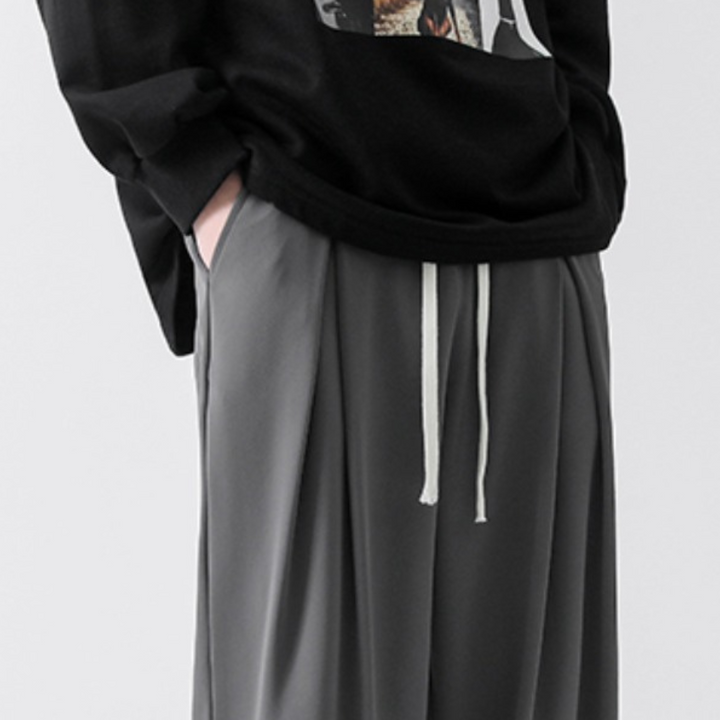 A person wears a black sweatshirt with a graphic design and grey streetwear joggers made from ice-silk fabric, featuring white drawstrings and an elastic waistband, with one hand in a pocket. The image is cropped at the shoulders and knees.