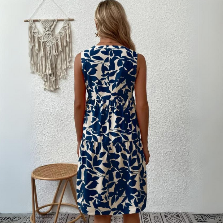 A woman with blonde hair stands facing a white textured wall, wearing a sleeveless dress in a blue and white leaf pattern perfect for a summer wardrobe. A woven wall hanging and a round wooden stool are seen beside her.