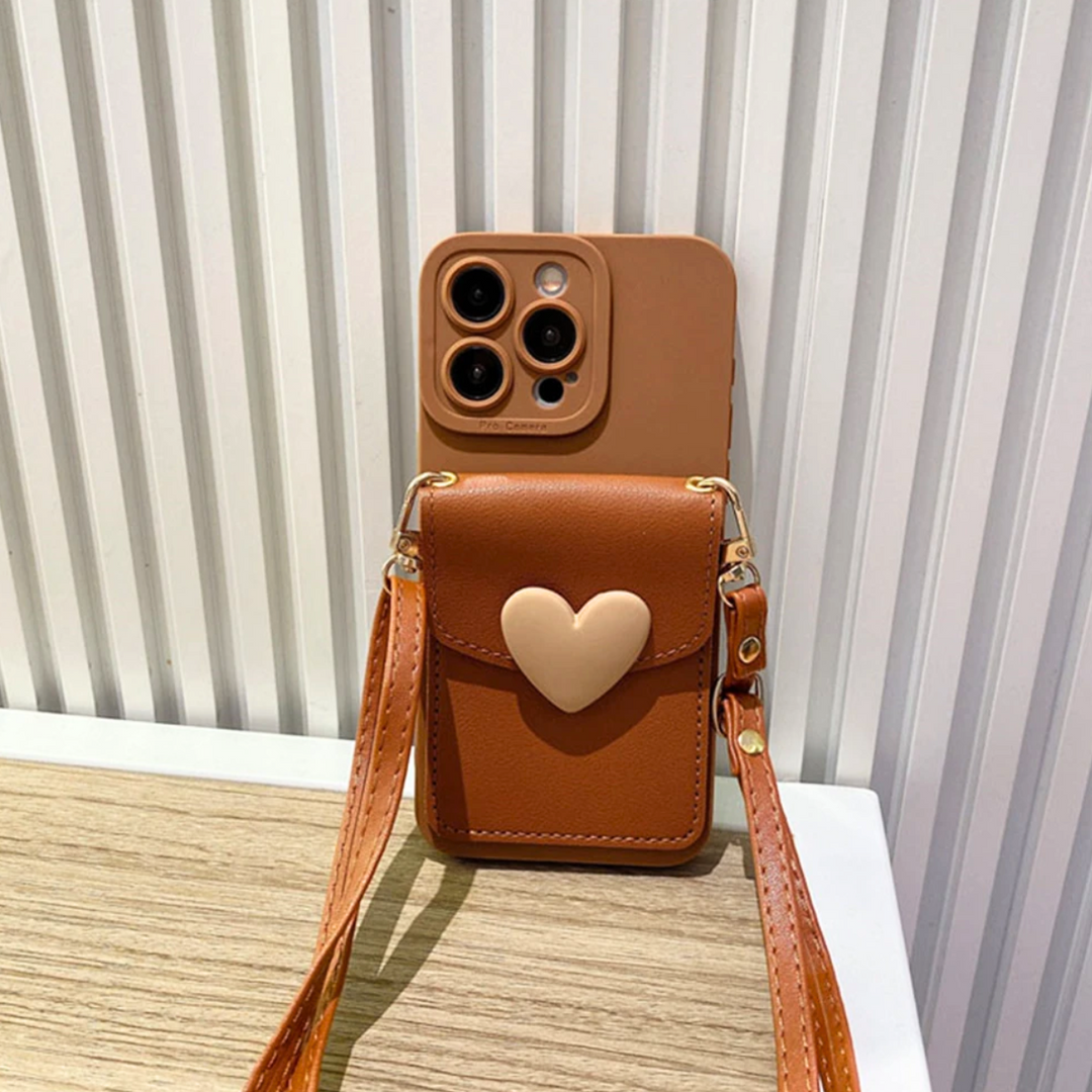 A chic phone case with a crossbody strap and a small heart-shaped pocket sits upright on a wooden surface, its brown design complemented by a beige heart, against a white, ridged wall.