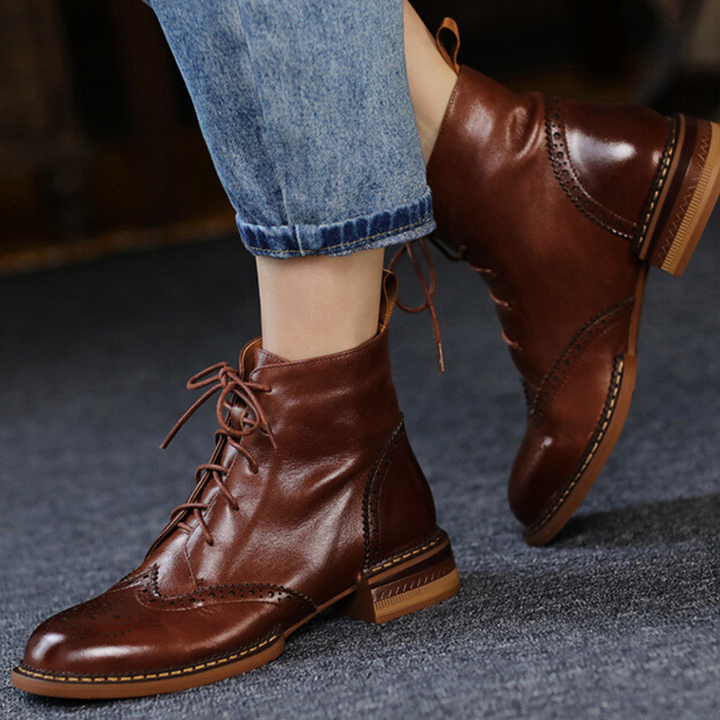 A stylish urban trendsetter wears cuffed blue jeans and brown leather brogue boots with decorative stitching, standing on a dark carpeted floor.