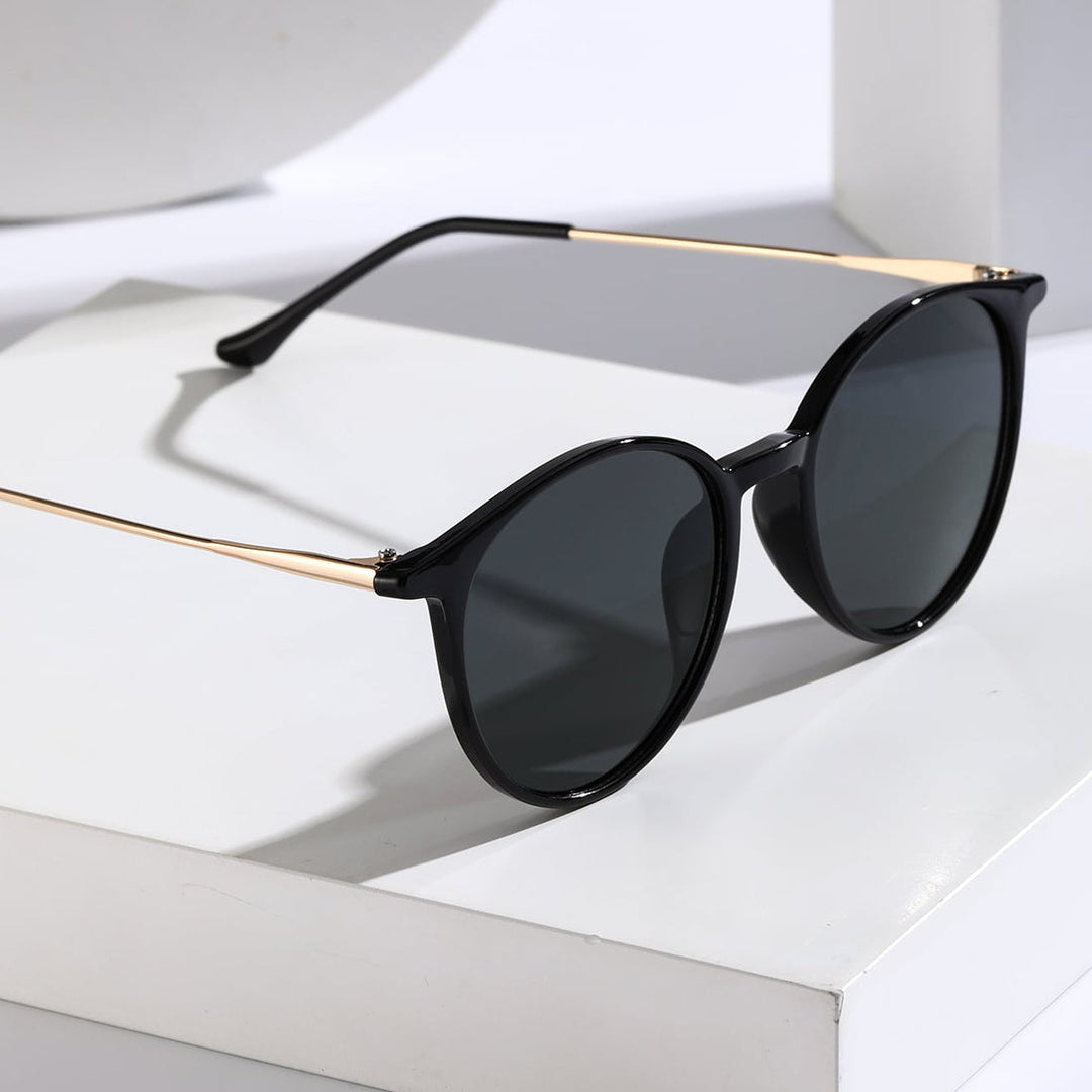 Black sunglasses with dark lenses and thin gold arms are displayed on a white surface, casting a shadow. The modern, round frame offers stylish design along with UV protection for your eyes.