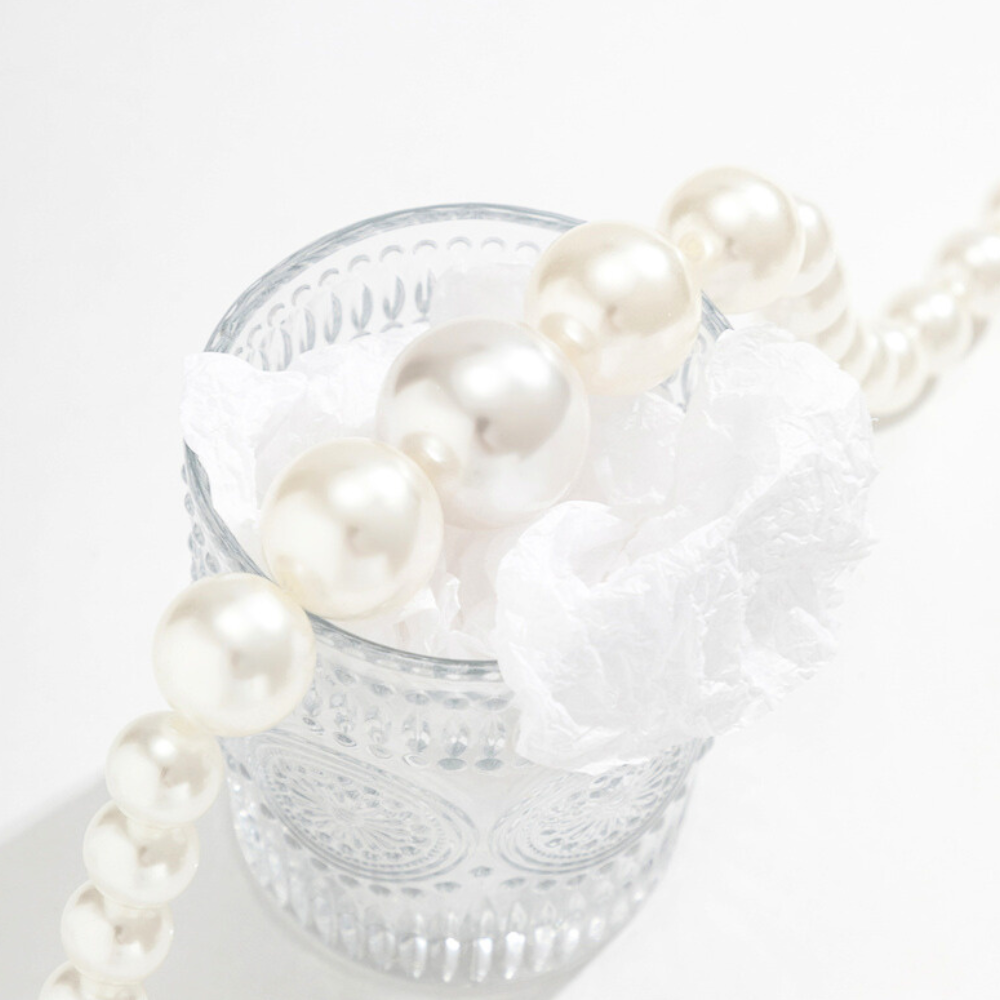 A string of white pearls, part of a chic pearl jewelry set, draped over the rim of a clear, textured glass cup filled with crumpled white tissue paper, set against a bright white background.