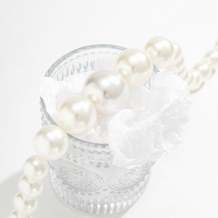A string of white pearls, part of a chic pearl jewelry set, draped over the rim of a clear, textured glass cup filled with crumpled white tissue paper, set against a bright white background.