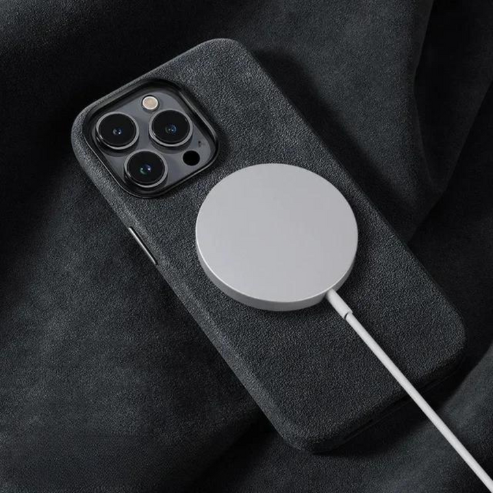 A smartphone with a dark, suede finish case rests on a soft, dark surface while a round, magnetic wireless charger is attached to its back.