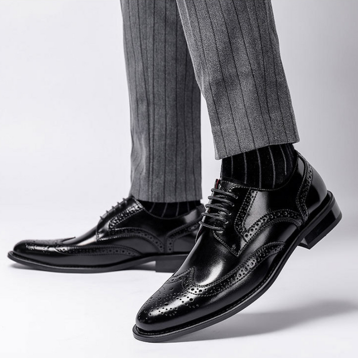 A person wearing premium leather brogue dress shoes with a cushioned insole, black socks, and grey pinstripe trousers stands on a white background.