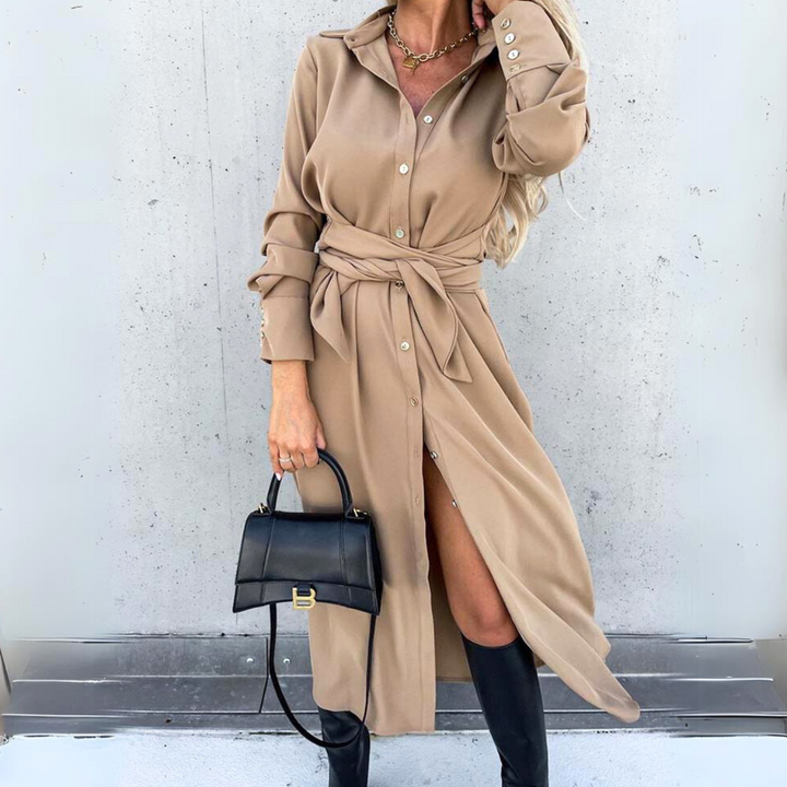 A woman wears a tan belted shirt dress styled with black knee-high boots and a small black handbag, exuding streetwear vibes. She stands against a light gray wall, her long blonde hair in loose waves.