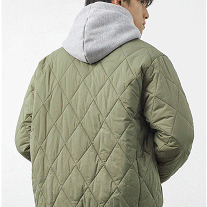A person is seen from behind wearing a light olive green, diamond-stitched padded jacket over a gray hooded sweatshirt. The background is plain and light-colored.