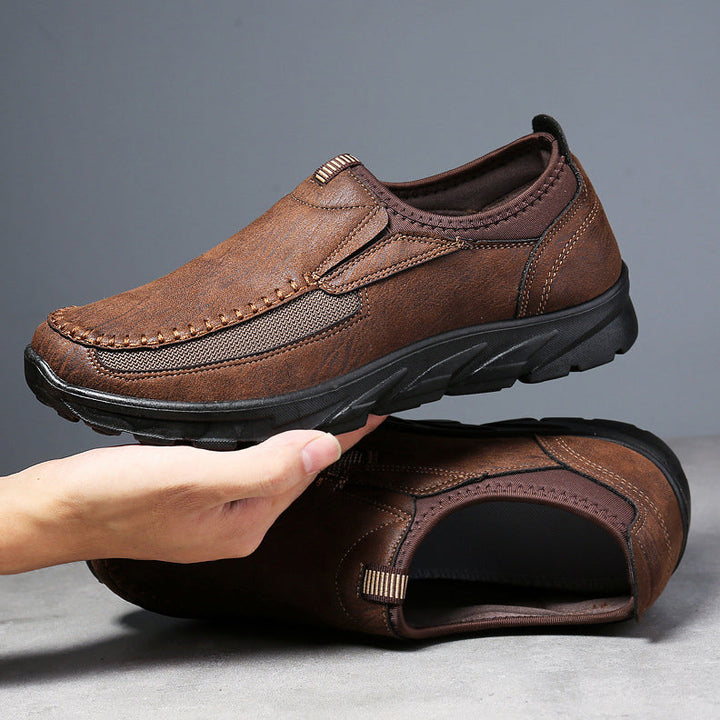 A hand holds up a brown Men Leather Casual Shoe with visible stitching and a black rubber sole, while another matching breathable loafer lies flat on a gray surface, both shoes showcased against a plain gray background.