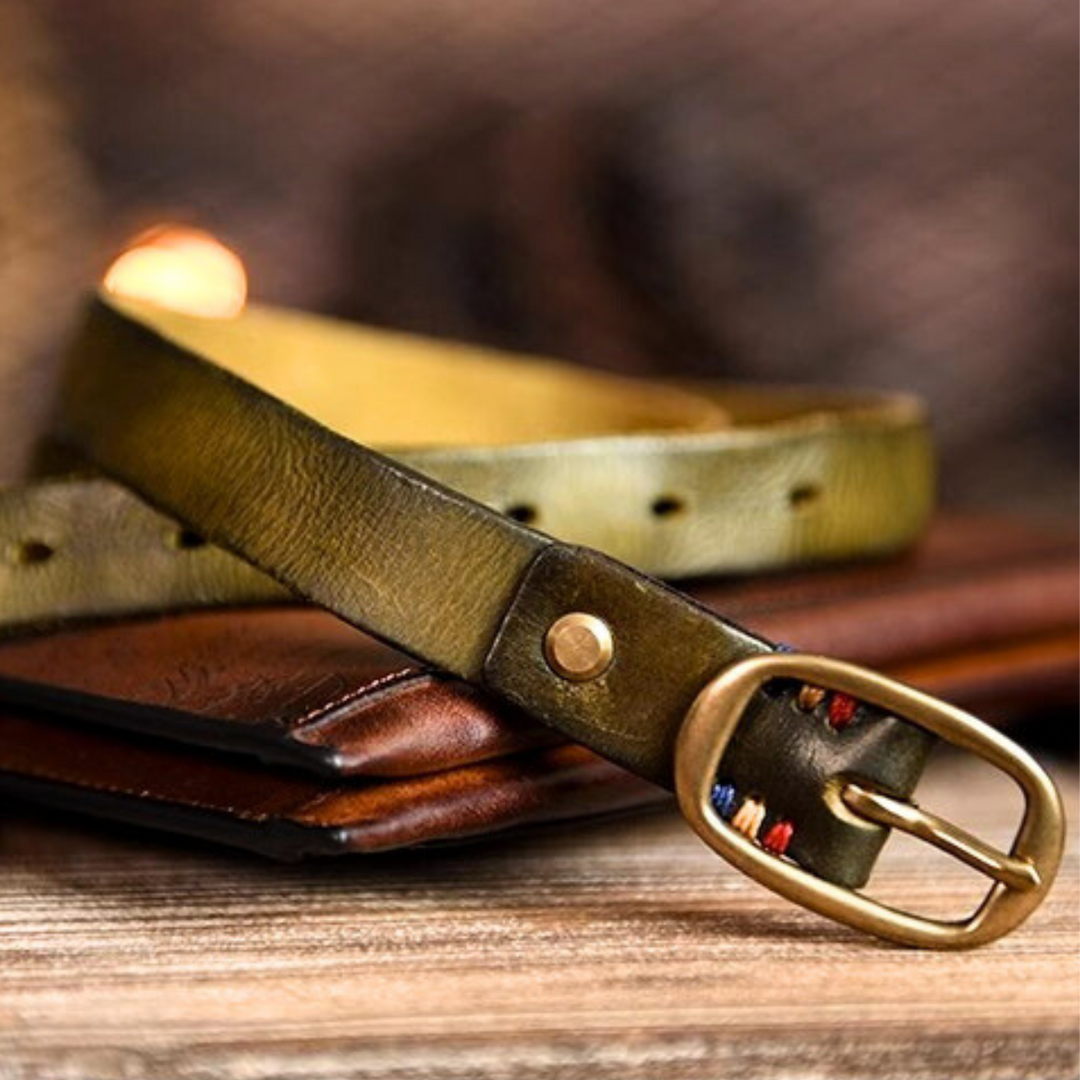 A close-up of an olive green leather belt with a brass buckle, resting on a brown leather wallet atop a wooden surface. Perfect for country fashion, the belt features decorative stitching near the buckle.