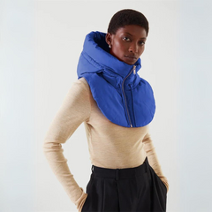 A person wearing a beige long-sleeve top, black pants, and a large blue puffer scarf made of durable material with a high collar and zipper, standing against a plain light background.