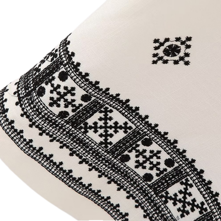 A close-up of white fabric on wide-leg trousers with intricate black geometric embroidery, featuring repeating cross and dot patterns along the edge and a smaller embroidered motif above.