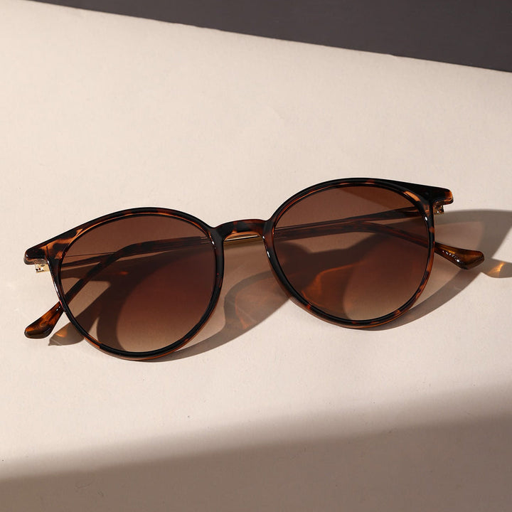 Tortoiseshell round sunglasses with brown tinted lenses offer UV protection, resting on a light-colored surface and casting a faint shadow.