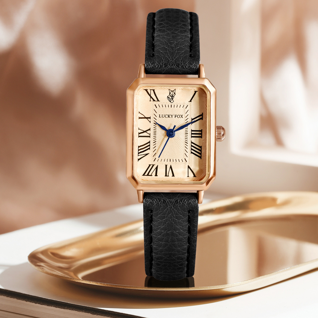 A rectangular face watch with a genuine leather strap, Roman numerals, and blue hands is displayed on a reflective gold tray against a softly blurred beige background.