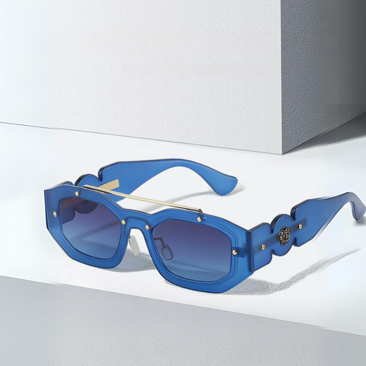 Blue, geometric sunglasses with gold accents and dark, scratch-resistant lenses rest on a white surface near a light gray wall, casting a soft shadow. The bold, modern design is complemented by reliable UV400 protection.