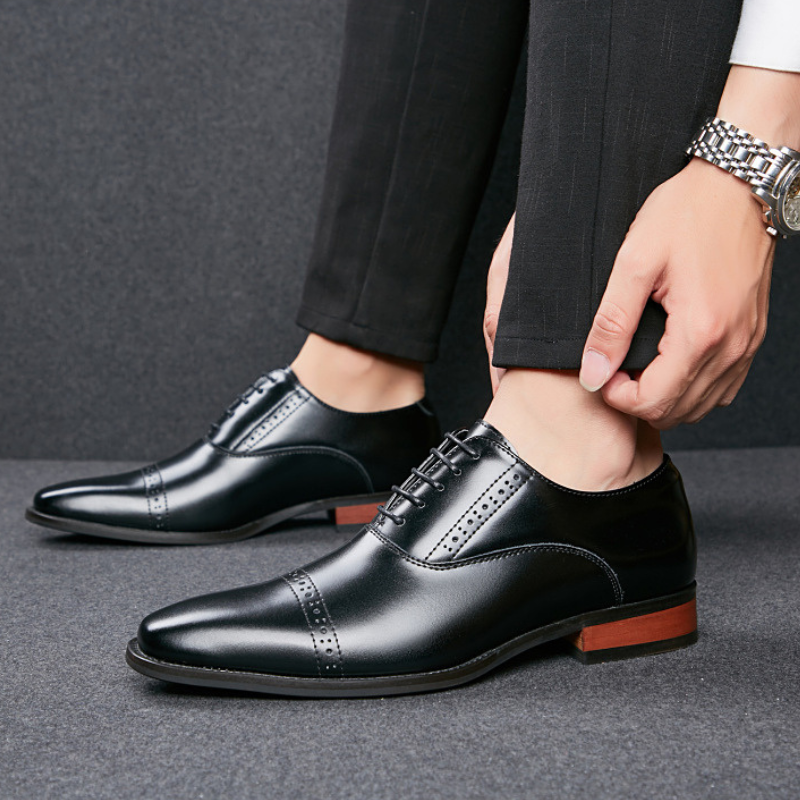 A person wearing black Italian leather dress shoes with brown soles and black trousers adjusts their pant leg, displaying a silver watch on their wrist. The background and floor are gray.