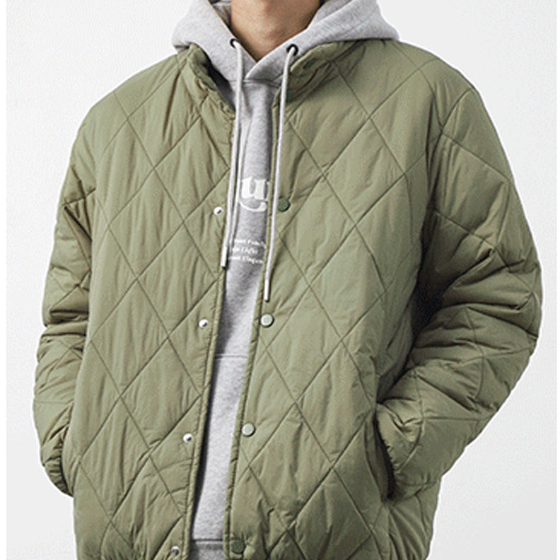 A person wearing a light olive padded jacket with a diamond-stitched pattern over a light gray hooded sweatshirt with white drawstrings, standing with hands in pockets against a plain background.