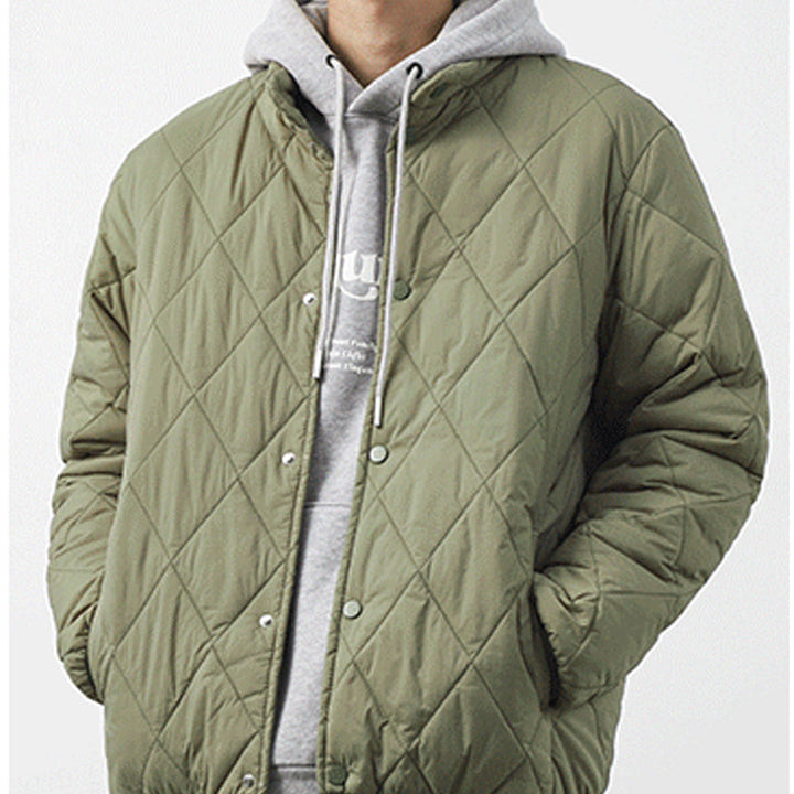 A person wearing a light olive padded jacket with a diamond-stitched pattern over a light gray hooded sweatshirt with white drawstrings, standing with hands in pockets against a plain background.
