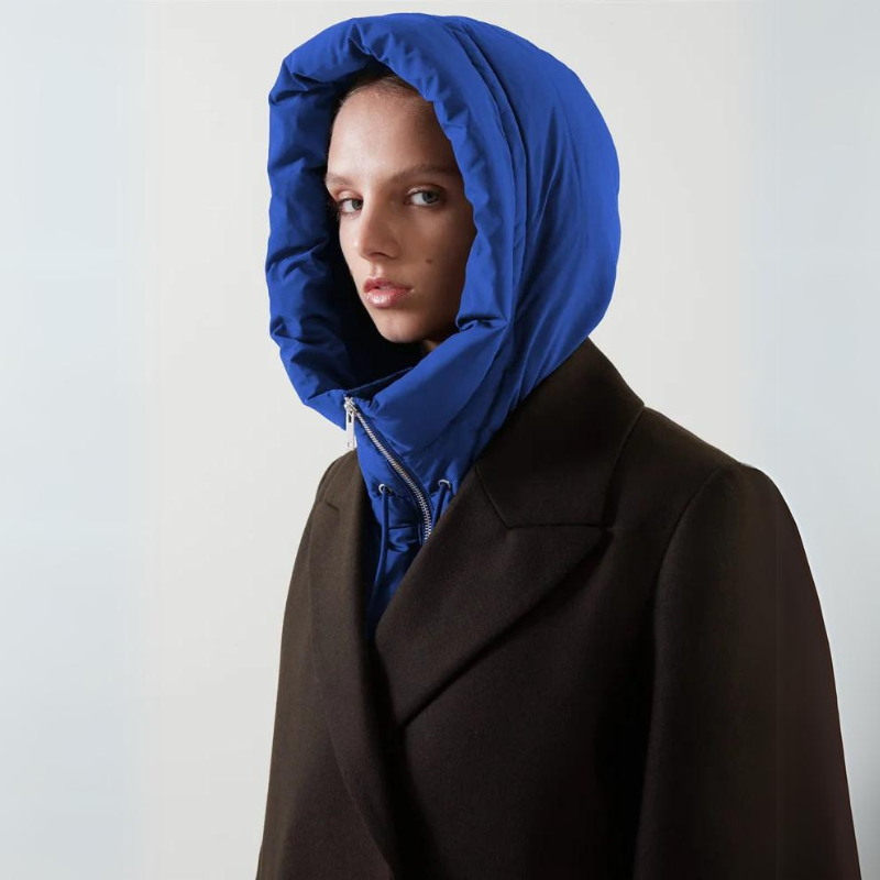 A person wearing a dark, lightweight coat with a bright blue, hooded design looks at the camera against a plain light background.