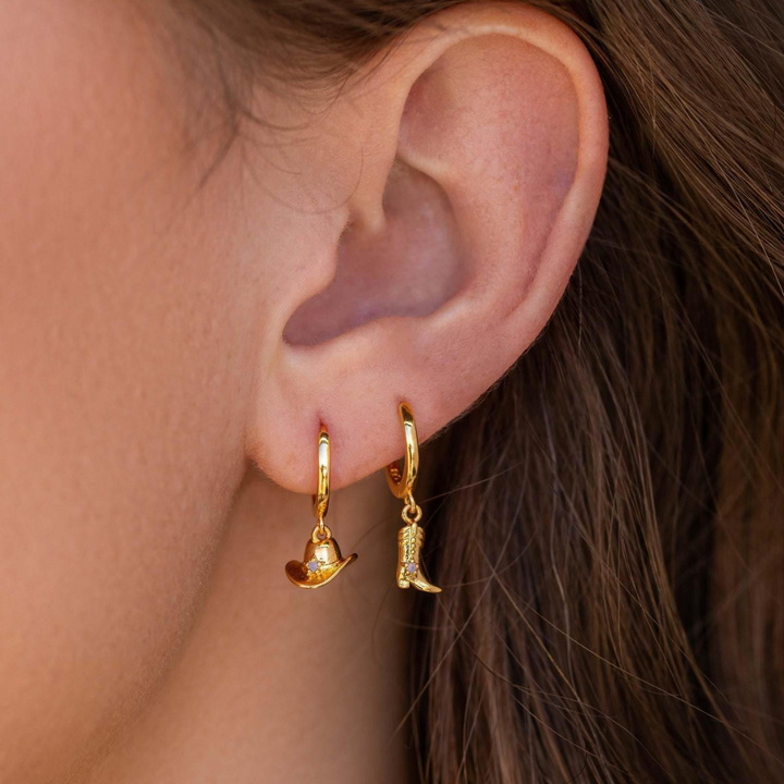 Close-up of a person’s ear wearing two small gold statement earrings: one with a dangling cowboy hat charm, the other with a cowboy boot. The person has light brown hair and fair skin, adding a playful urban chic vibe.