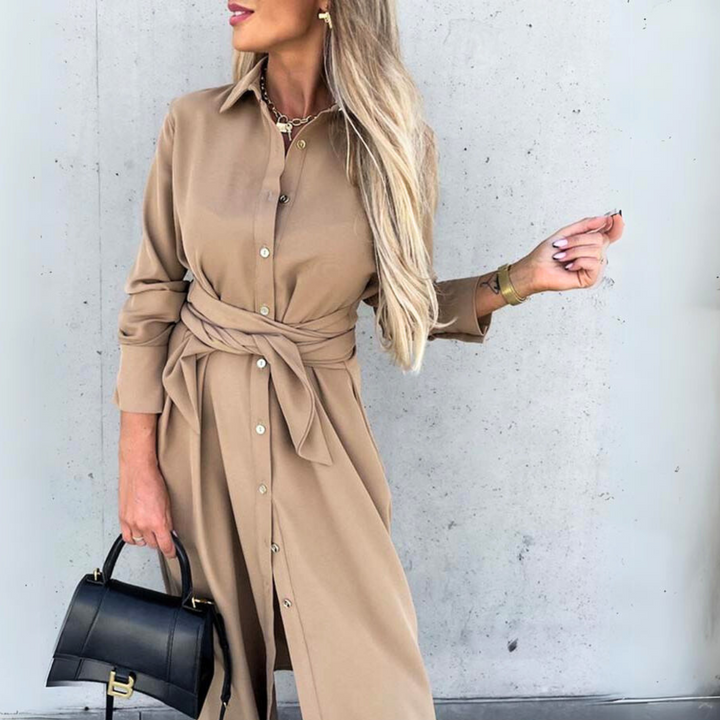A woman wearing a tan belted shirt dress stands against a light gray wall, holding a black handbag. Her long blonde hair is styled down, and she accessorizes with a gold necklace and bracelet for effortless streetwear appeal.