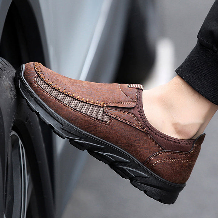 A person wearing brown breathable loafers with black rubber soles rests their foot against a car tire, showcasing the comfortable fit and stitching details of these Men Leather Casual Shoes.